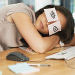 Une femme en train de dormir sur son bureau devant son ordinateur avec des post-it sur les yeux