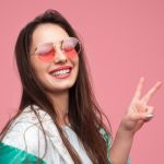 Une jeune femme sur fond rose en lunettes rose fait un signe peace des doigts