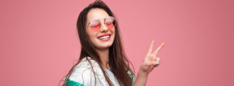 Une jeune femme sur fond rose en lunettes rose fait un signe peace des doigts