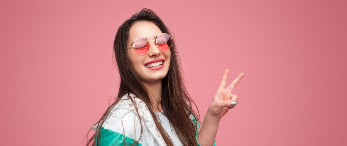 Une jeune femme sur fond rose en lunettes rose fait un signe peace des doigts