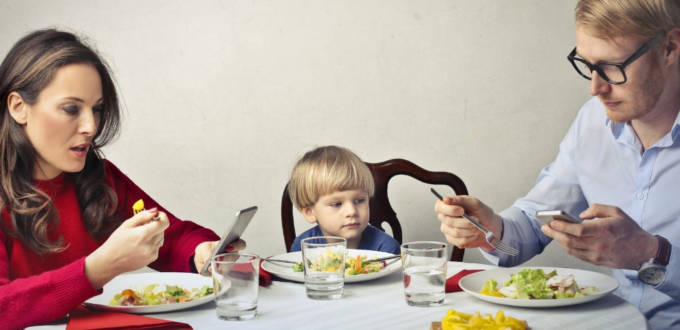 Un homme et une femme à table en train de regarder leur smartphone pendant qu'un enfant est seul au milieu