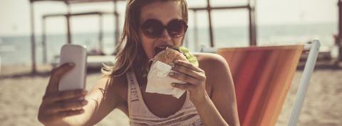 Regarder la junk food sur Insta fait grossir Une femme à la plage qui prend un selfie en mangeant un burger