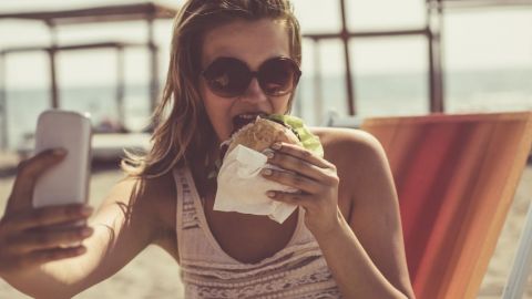 Une femme à la plage qui prend un selfie en mangeant un burger