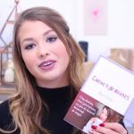 une jeune fille montre un livre devant un fond rose.