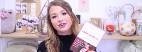 EnjoyPhoenix présente son nouvel ouvrage une jeune fille montre un livre devant un fond rose.