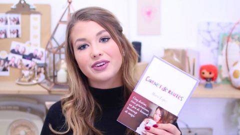 EnjoyPhoenix présente son nouvel ouvrage une jeune fille montre un livre devant un fond rose.