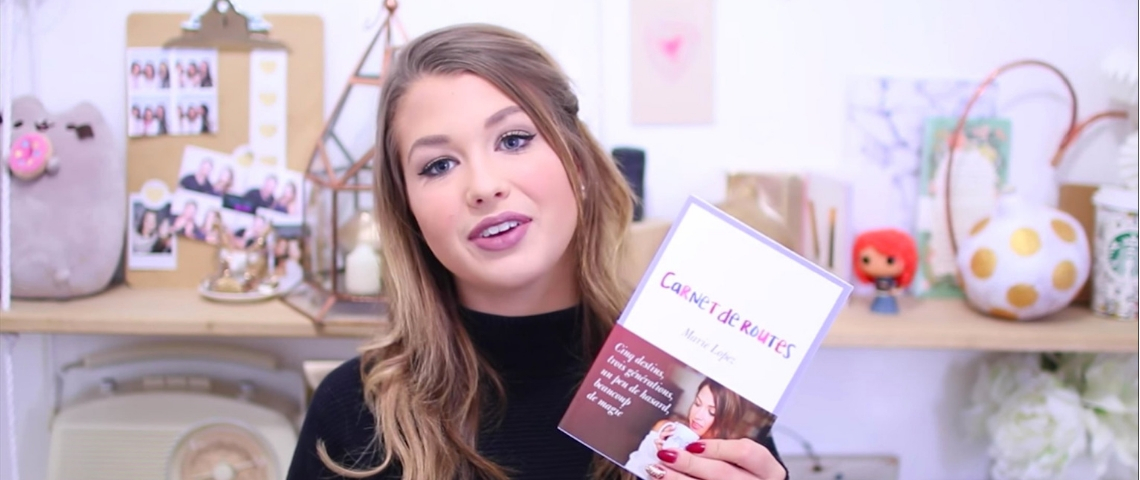 EnjoyPhoenix présente son nouvel ouvrage une jeune fille montre un livre devant un fond rose.