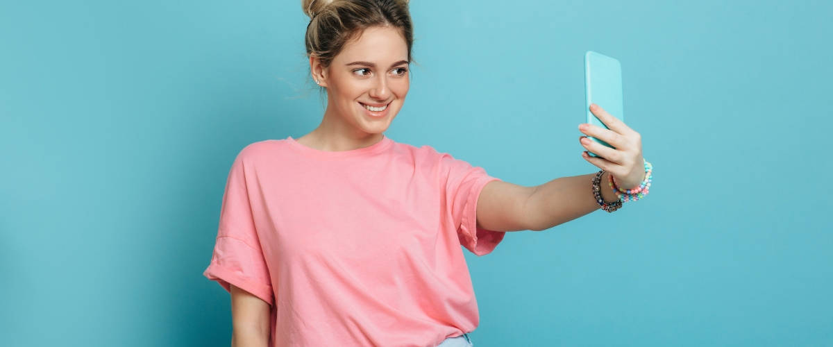 Social media : des bonnes pratiques aux mauvaises habitudes Une femme avec un t-shirt rose qui prend un selfie devant un fond bleu