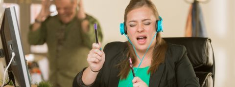 Comment les salariés vivent-ils le bruit au bureau ? Une jeune femme en train d'écouter de la musique bruyamment à son bureau et un homme énervé en arrière-plan