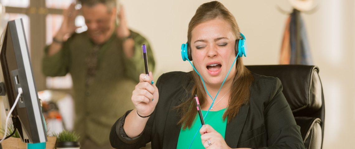 Comment les salariés vivent-ils le bruit au bureau ? Une jeune femme en train d'écouter de la musique bruyamment à son bureau et un homme énervé en arrière-plan