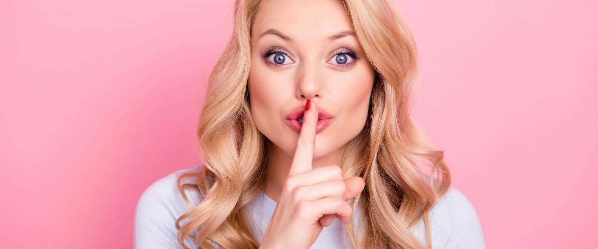 Les chiffres de l'infidélité féminine en 2019 Une femme blonde qui fait chut avec son doigt devant un fond rose