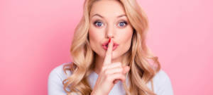 Une femme blonde qui fait chut avec son doigt devant un fond rose