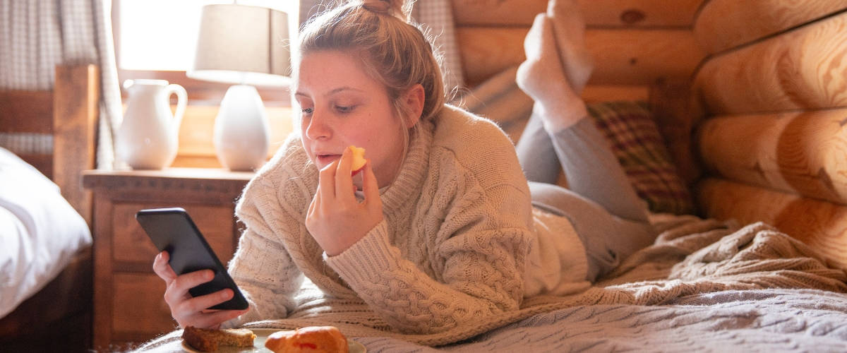 Quel est l'impact des écrans sur nos repas ? Une fille blonde qui mange allongée sur son lit en regardant son téléphone