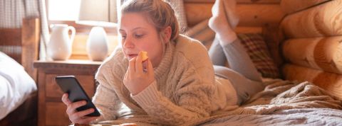 Quel est l'impact des écrans sur nos repas ? Une fille blonde qui mange allongée sur son lit en regardant son téléphone