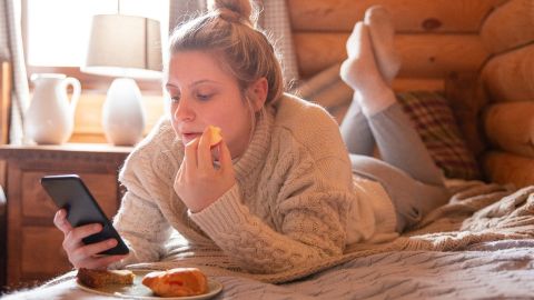 Une fille blonde qui mange allongée sur son lit en regardant son téléphone