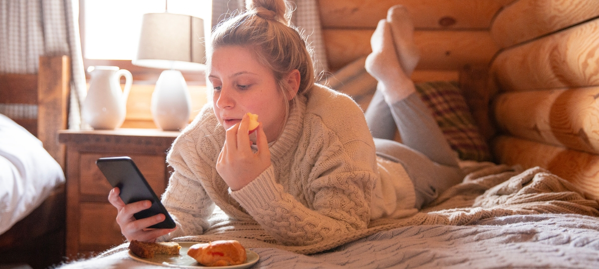 Quel est l'impact des écrans sur nos repas ? Une fille blonde qui mange allongée sur son lit en regardant son téléphone