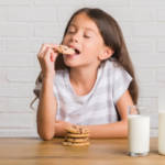 Un jeune enfant aux cheveux longs en train de manger un cookie trempé dans un verre de lait