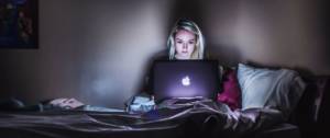 Une jeune femme blonde regarde l'écran de son Macbook sur son lit