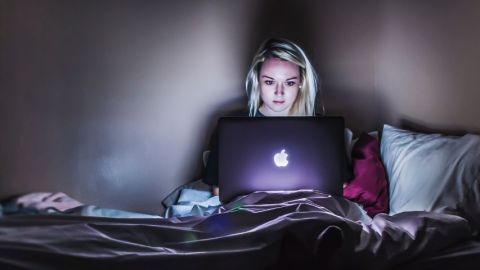 Une jeune femme blonde regarde l'écran de son Macbook sur son lit
