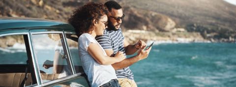 5 applis pour des vacances insolites Couple adossé contre voiture vintage à la mer, regardent un smartphone