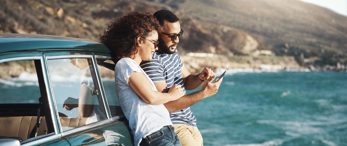 5 applis pour des vacances insolites Couple adossé contre voiture vintage à la mer, regardent un smartphone