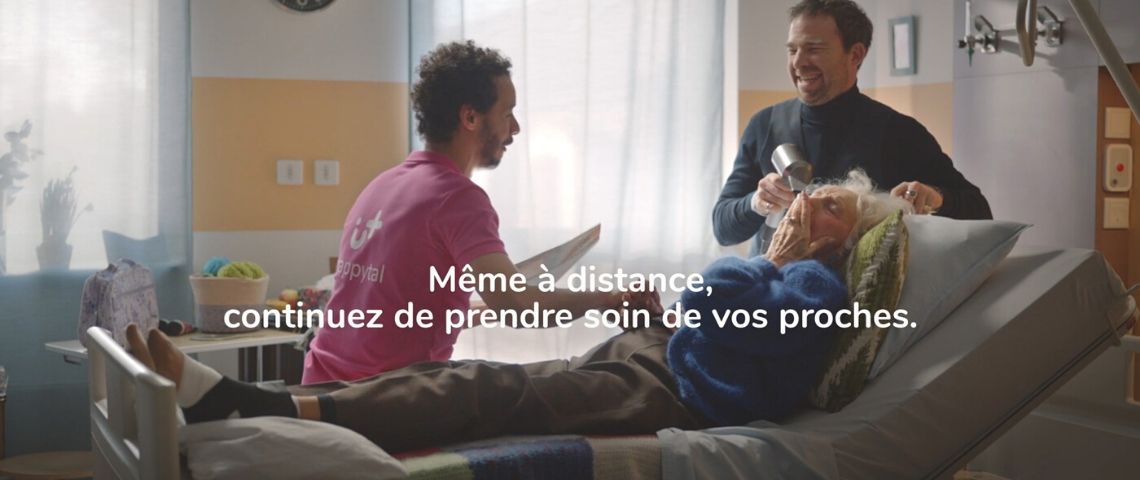 Avec Happytal et Buzzman, prenez soin de vos proches même à distance Homme assis sur un lit d'hopital en train de converser avec une femme agée pendant qu'elle se fait coiffer, le tout en riant