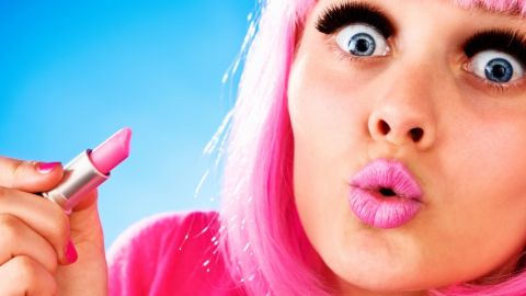 Une femme très maquillée portant une perruque rose et tenant un tube de rose à lèvres