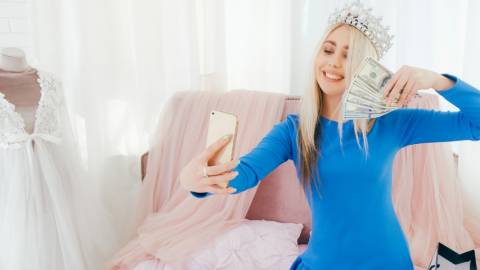 une jeune fille blonde avec une couronne sur la tête se prend en photo avec une liasse de billets dans la main.