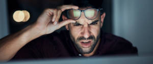 un homme fronce les sourcils et lève ses lunettes en regardant un écran.