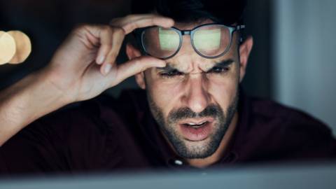 un homme fronce les sourcils et lève ses lunettes en regardant un écran.
