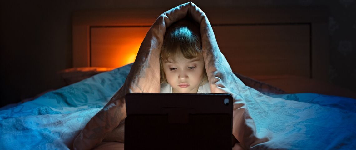 Comment gérer les écrans pendant le confinement ? Une petite fille qui joue sur un ordinateur cachée sous sa couette