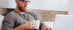 Un homme dans son lit en train de boire un café et d'utiliser son ordinateur