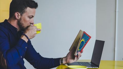 Un homme lit un livre devant son ordinateur portable