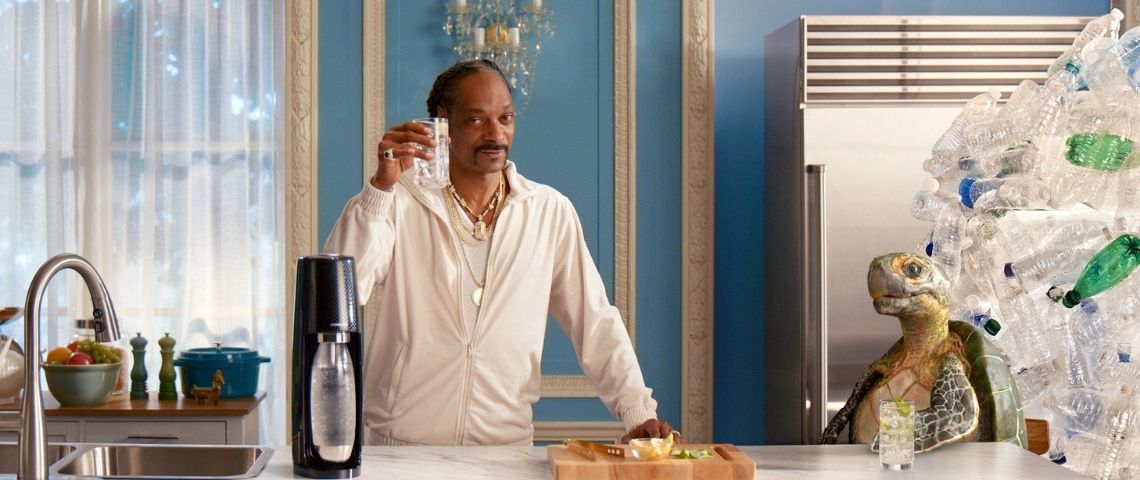 A l’occasion des fêtes de fin d’année Sodastream et Snoop Dogg vous souhaitent un joyeux noël 2020 Snoop Dogg, un verre de sodastream à la main avec une tortue et des déchets de bouteilles plastiques derrière elle