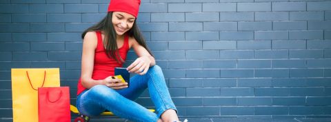 Les ados n'ont aucun tabou avec l'argent, et les fintech en profitent Adolescente assise par terre regarde son smartphone, avec une carte bancaire dans une main et des sacs de shopping à côté d'elle