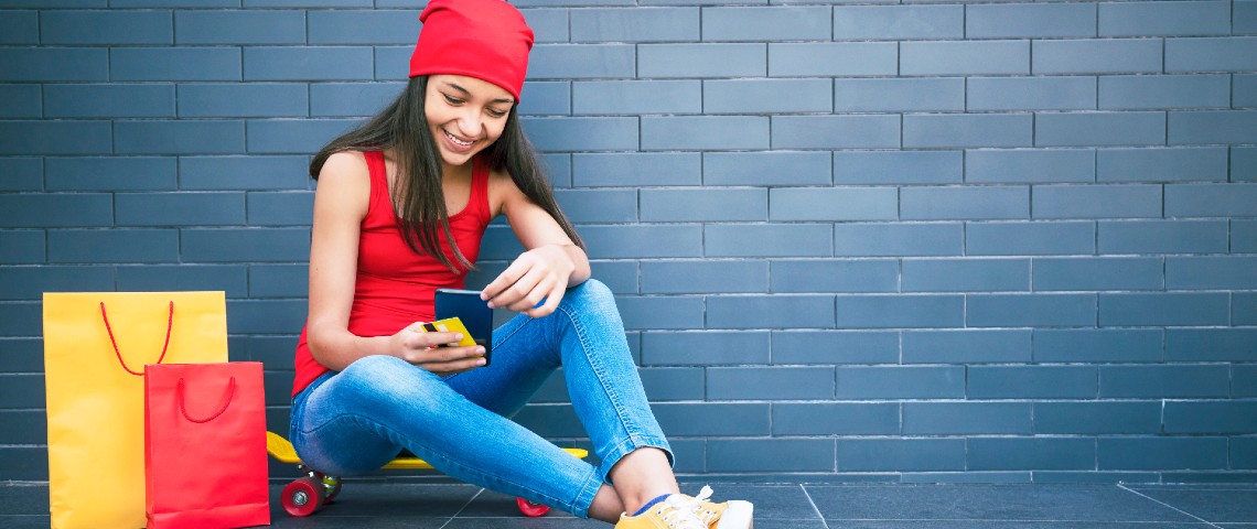 Les ados n'ont aucun tabou avec l'argent, et les fintech en profitent Adolescente assise par terre regarde son smartphone, avec une carte bancaire dans une main et des sacs de shopping à côté d'elle
