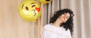 Une femme brune tenant dans la main un ballon en forme de smiley qui envoie un bisou coeur