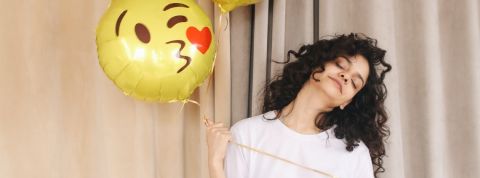 Comment se remonter le moral pendant le confinement ? Une femme brune tenant dans la main un ballon en forme de smiley qui envoie un bisou coeur
