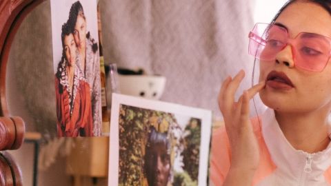 Une femme en train de se maquiller devant un miroir, portant un turban et des lunettes roses