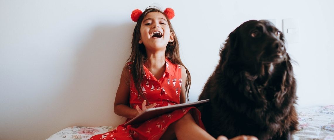 Keeku, une plateforme gratuite, réunissant 1500 podacsts pour les enfants de 0 à 12 ans Petite riant avec une tablette dans les mains, assise sur son lit, accompagnée de son chien