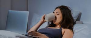 Une femme en pyjama dans son lit devant son ordinateur portable en train de boire une tasse de café