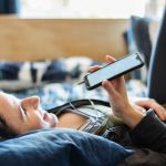 Une femme sur son lit parle dans le micro de son smartphone et porte un casque