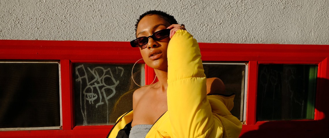 Comment l'appli Depop repère les tendances avant tout le monde Une femme pose avec des lunettes rectangulaire et une grosse doudoune jaune