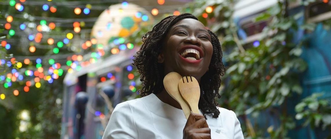 Meet My Mama un modèle entrepreneurial hybride pour aider des femmes réfugiées ou immigrées à s'émanciper financièrement Femme noire avec des couverts à salade en bois, riant aux éclats