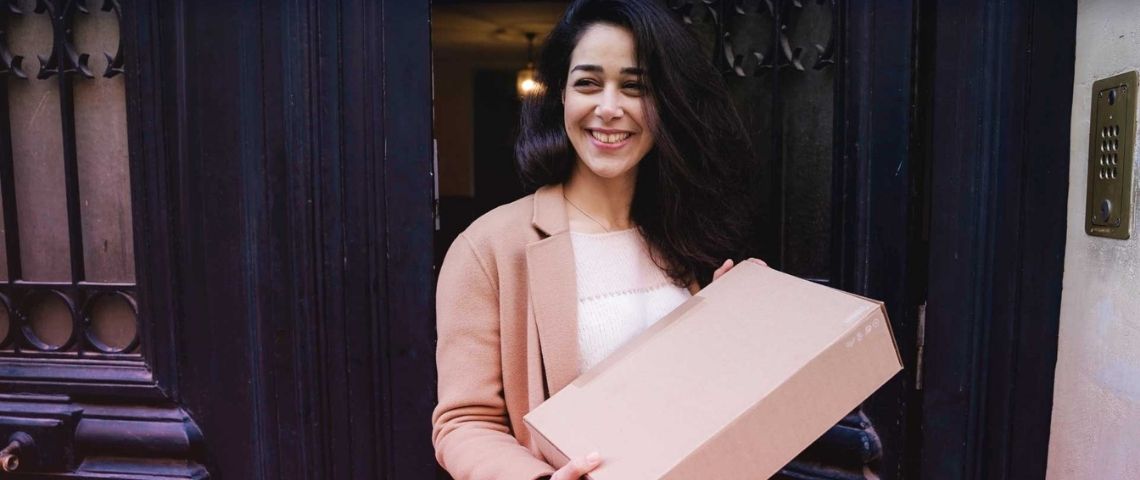 Pickme est la première plateforme de livraison collaborative à transformer les voisins en un réseau de points-relais de proximité. Jeune femme, devant la porte de son immeuble, tenant un colis dans ses mains