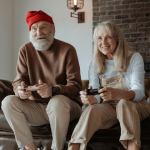 Un couple de personnes âgées joue aux jeux vidéos sur le canapé de leur salon. Ils semblent regarder vers l'écran de télévision et ont des manettes dans les mains. Ils ont l'air enthousiasmés par leur partie et sourient. Le grand père a un bonnet, une grosse barbe, un pull et un pantalon. La femme porte un tee-shirt à manches longues et une salopette.