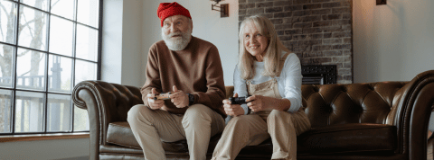 Un couple de personnes âgées joue aux jeux vidéos sur le canapé de leur salon. Ils semblent regarder vers l'écran de télévision et ont des manettes dans les mains. Ils ont l'air enthousiasmés par leur partie et sourient. Le grand père a un bonnet, une grosse barbe, un pull et un pantalon. La femme porte un tee-shirt à manches longues et une salopette.