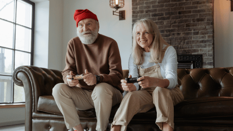 Un couple de personnes âgées joue aux jeux vidéos sur le canapé de leur salon. Ils semblent regarder vers l'écran de télévision et ont des manettes dans les mains. Ils ont l'air enthousiasmés par leur partie et sourient. Le grand père a un bonnet, une grosse barbe, un pull et un pantalon. La femme porte un tee-shirt à manches longues et une salopette.