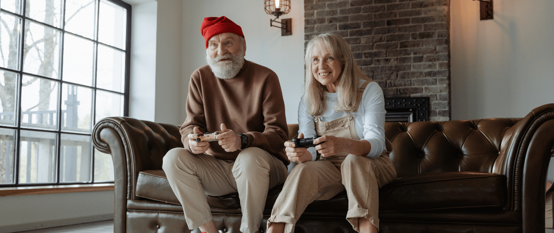Qui sont les papis gamers ? Un couple de personnes âgées joue aux jeux vidéos sur le canapé de leur salon. Ils semblent regarder vers l'écran de télévision et ont des manettes dans les mains. Ils ont l'air enthousiasmés par leur partie et sourient. Le grand père a un bonnet, une grosse barbe, un pull et un pantalon. La femme porte un tee-shirt à manches longues et une salopette.