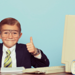 Un enfant en costume noir assis à un bureau le pouce en l'air